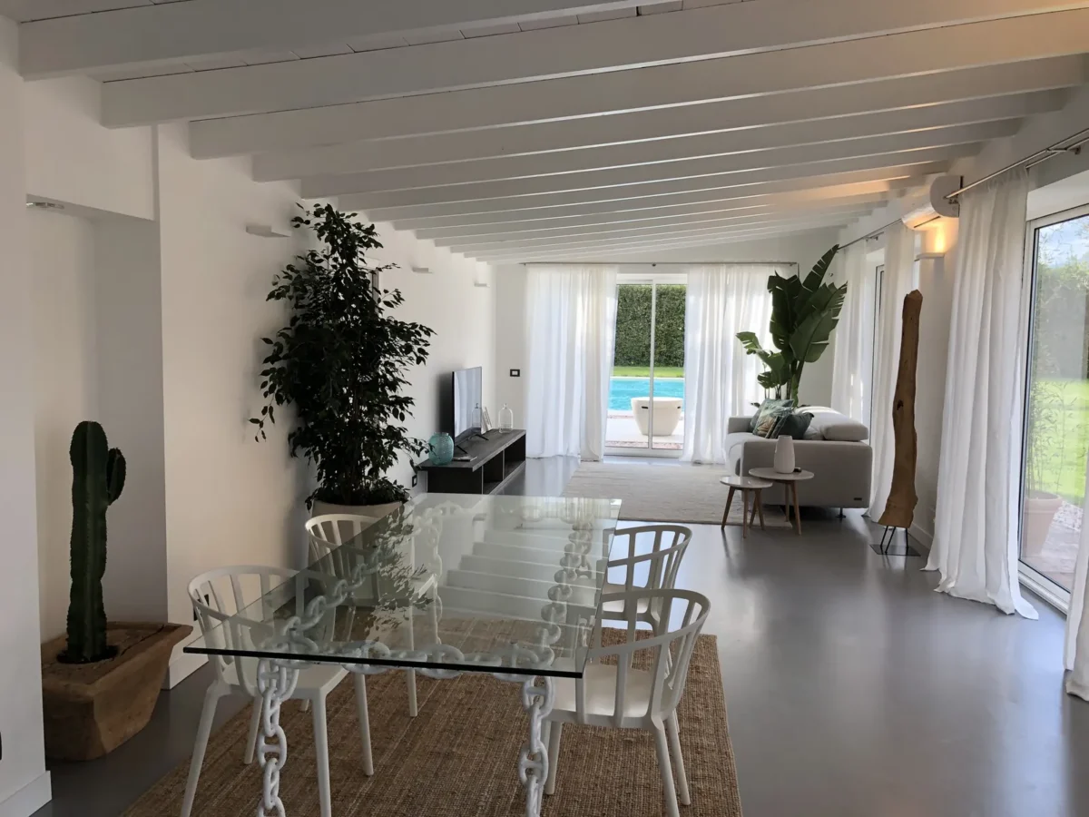 Open-plan living and dining area of the BELLA apartment with white beams, linen curtains and the pool through the glass door